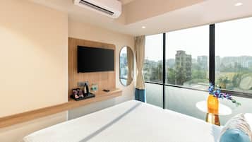 Habitación de hotel con una cama grande, un televisor montado, una mesita de noche con un florero y vistas a la ciudad a través de amplios ventanales.