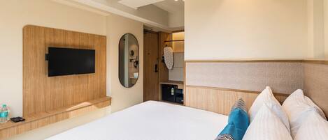 Habitación de hotel con una cama grande, televisor montado, cabecera de madera, mesita de noche con una botella y control remoto, y aire acondicionado instalado en el techo.