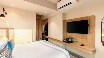 A modern hotel room with a bed, a mounted TV, a wooden shelf with toiletries, and an air conditioning unit.