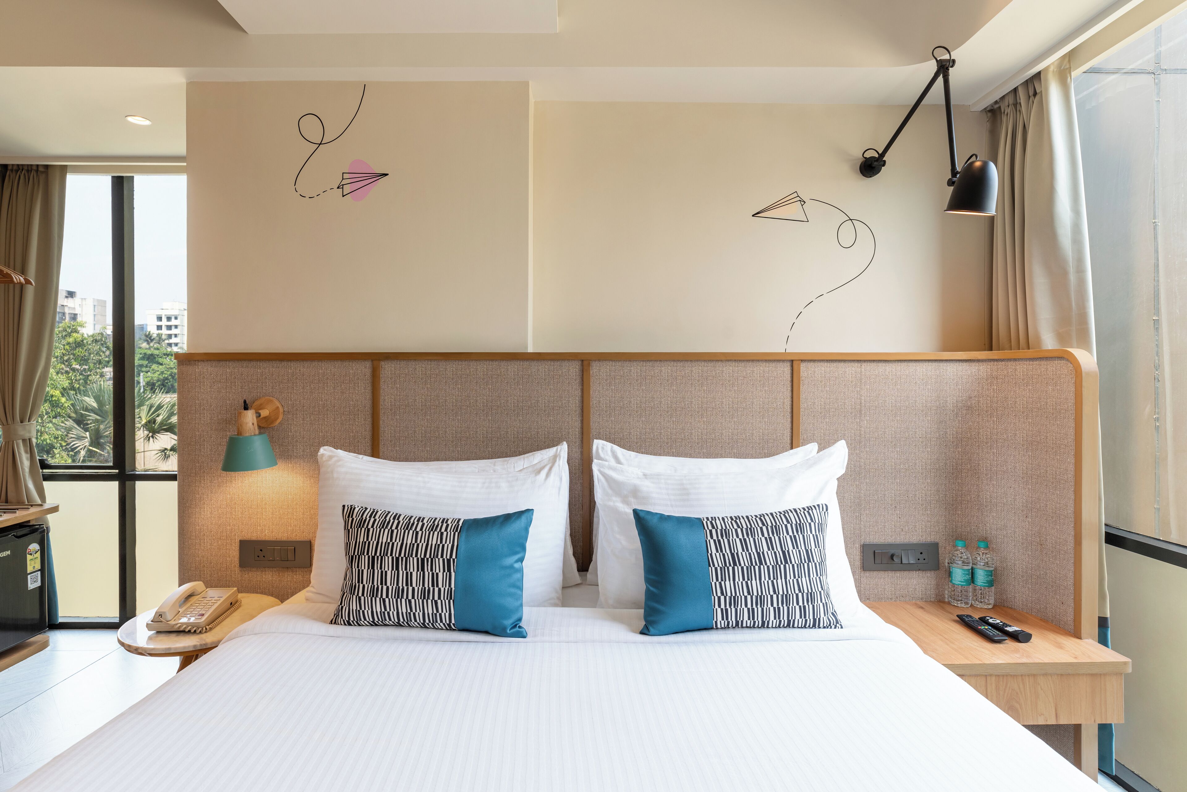 A hotel room with a large bed, two bedside tables, a nightstand, a lamp, a phone, and a view of the city through the window.