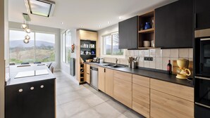 A modern kitchen with a large island, wooden cabinets, a built-in oven, and a view of the outdoors. - Architect-designed house with swimming pool 10 minutes from St Jean de Luz (Urrugne)