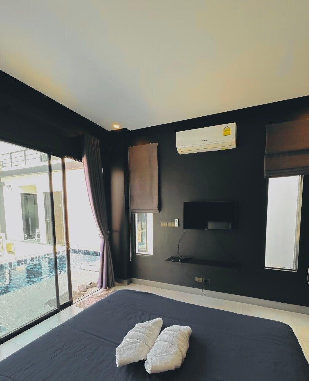 A modern hotel room with a large bed, a flat-screen TV, an air conditioning unit, and a view of a swimming pool.