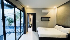 Une chambre d’hôtel moderne dotée d’une grande porte-fenêtre coulissante donnant sur un balcon, d’un lit avec du linge de maison blanc, d’un climatiseur fixé au mur et d’une petite étagère.