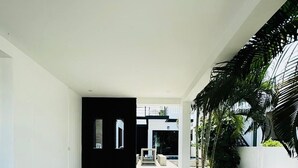 Une entrée moderne avec une porte noire, un chemin en béton menant à une zone abritée et un petit jardin agrémenté de palmiers.