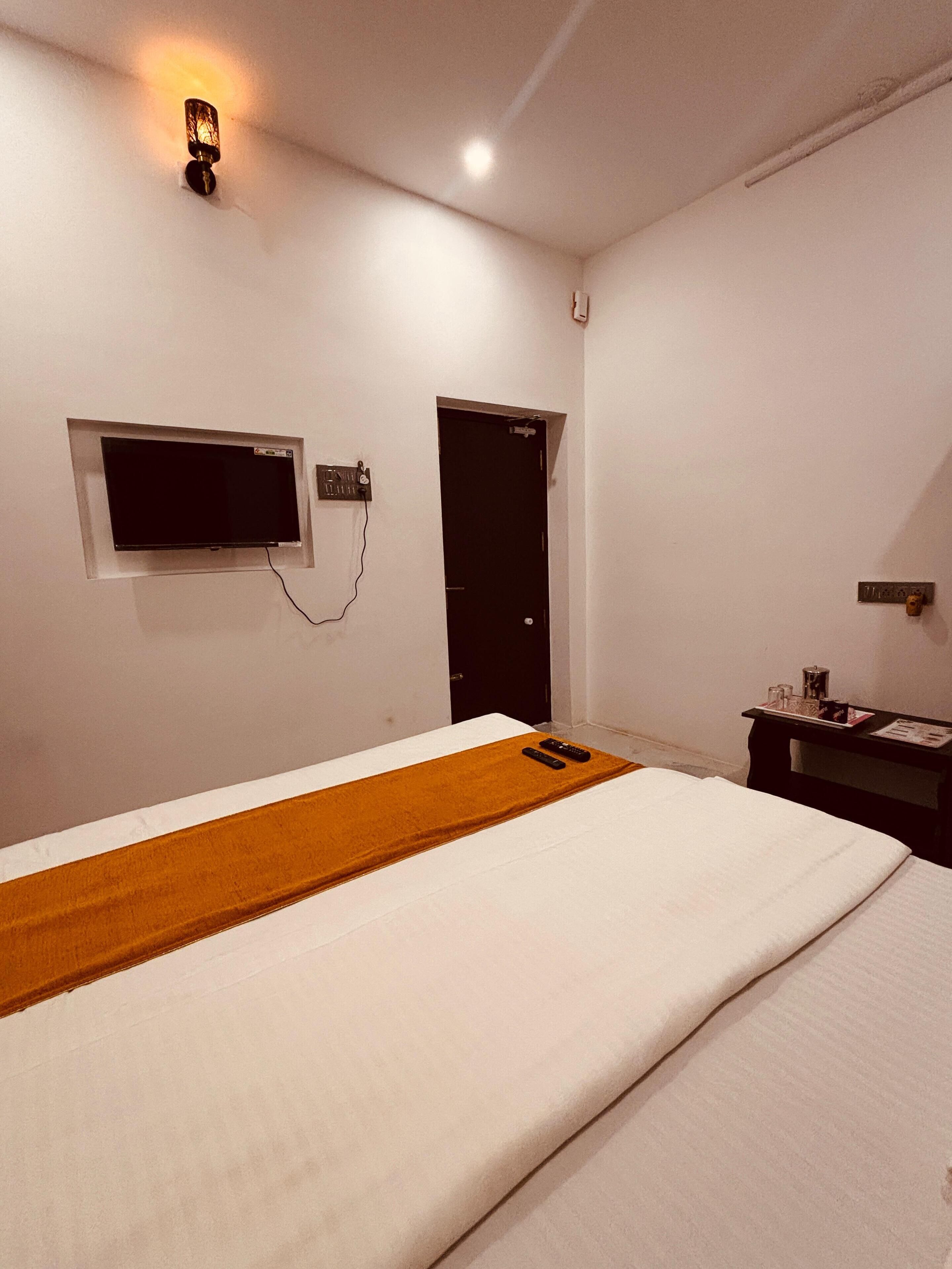 Una habitación de hotel con una cama, un televisor en la pared, una puerta y una mesita con objetos encima.