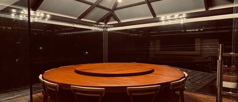 Une table ronde en bois, entourée de six chaises, dans un intérieur moderne agrémenté d’un plafond en verre aux motifs géométriques et d’un lustre.