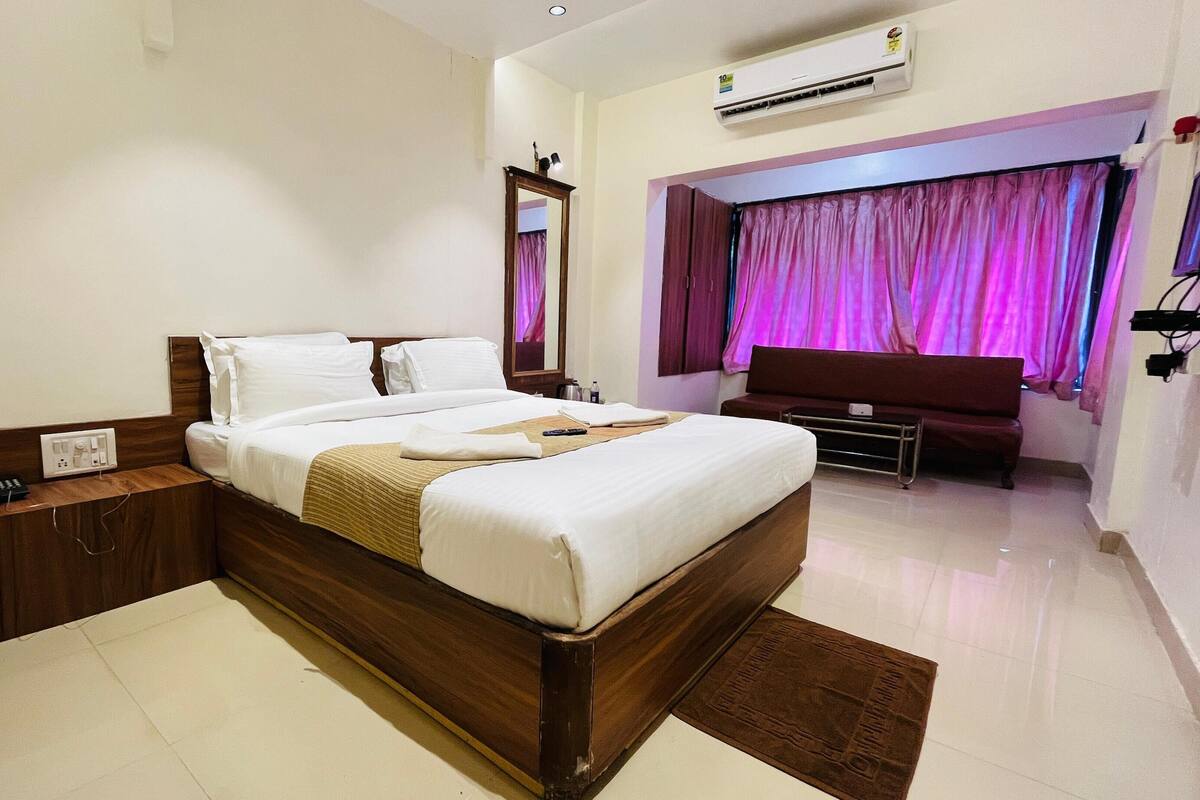 A hotel room with a large bed, a wooden nightstand, a desk, a mirror, a TV, and a window with curtains.