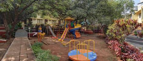A playground with various play structures, a paved walkway, and surrounding greenery.