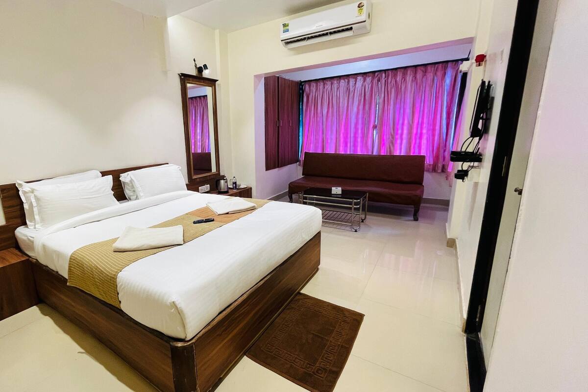 A hotel room with a large bed, bedside tables, a desk, a chair, a sofa, and a bathroom with a mirror and curtains.