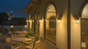 Un espace extérieur aménagé en terrasse, avec des tables et des chaises, un coin salon couvert avec des arcades, et une vue sur l’eau en arrière-plan.