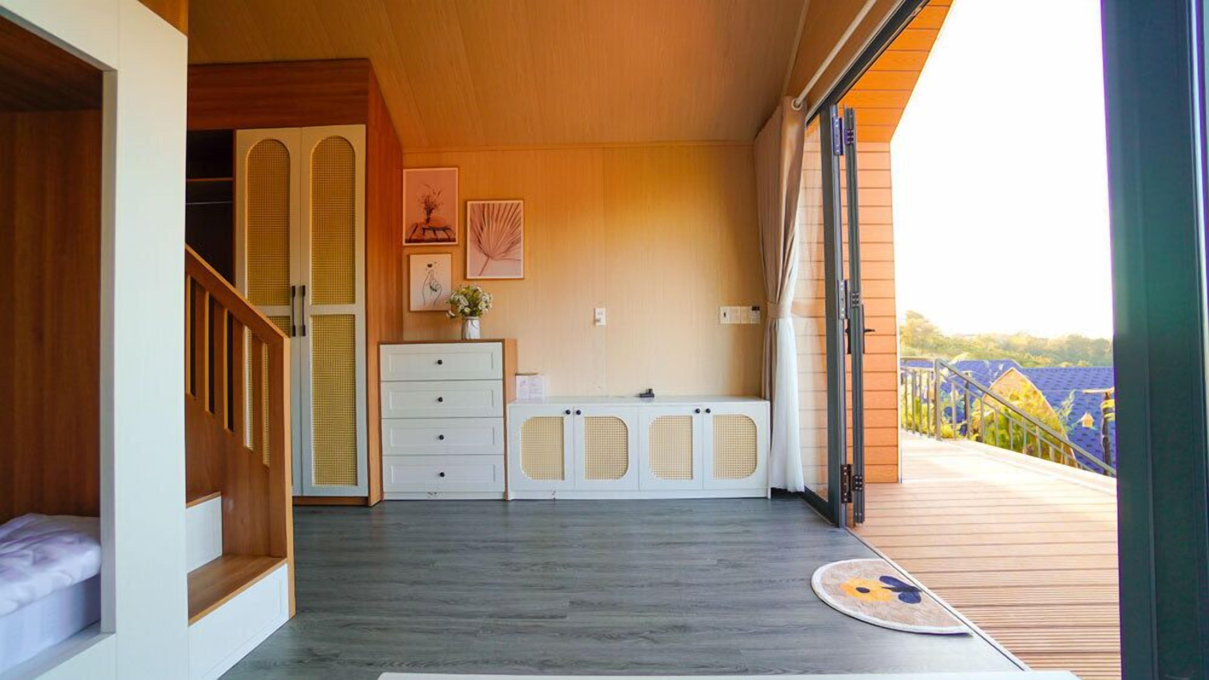 A room with a wooden floor, a white dresser, a bed with a white headboard, a staircase, and a balcony with a view.