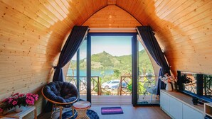 A cozy room with a wooden ceiling, a black chair, a small table, a potted plant, and a view of a lake and cars outside. - EVA VILLAGE (Dong Gia Nghia)