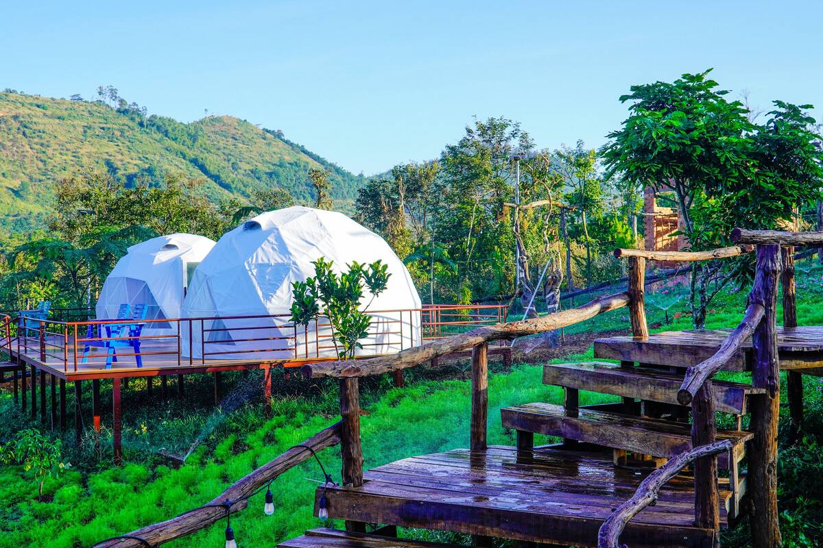 Two dome-shaped yurts connected by a wooden bridge, set in a lush green forest with a mountain in the background.