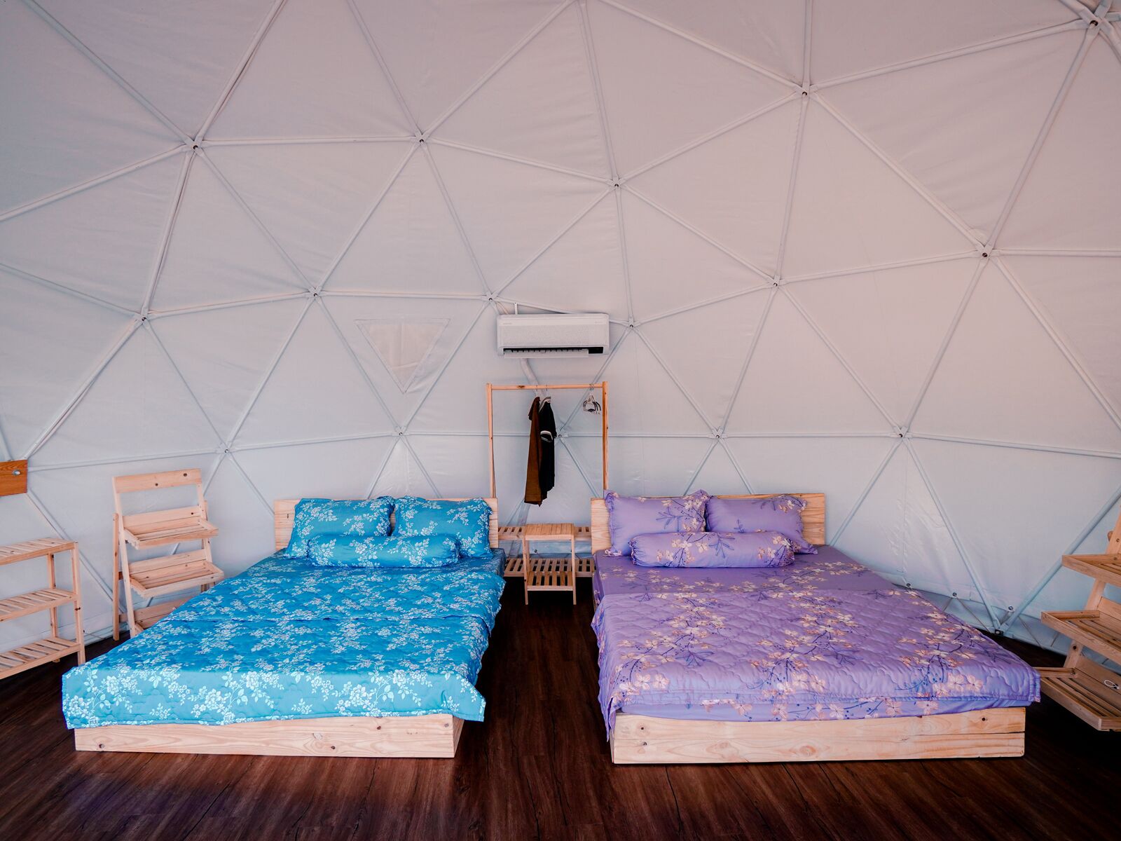 A tented room with two wooden beds, each with floral bedding, and a central air conditioning unit.