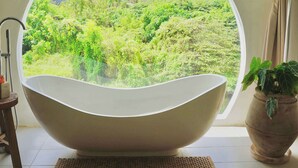 Un baño moderno con una bañera ovalada grande, vistas a zonas verdes exteriores y una planta en maceta sobre el alféizar.