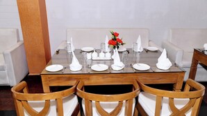 A dining table set with white plates, glasses, and napkins, surrounded by wooden chairs with white cushions. - Hotel Pardesi Cardo (Saharsa)