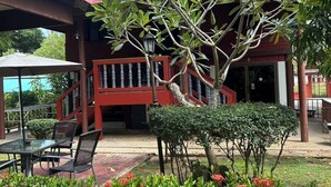 A covered outdoor seating area with a red-roofed building, a staircase, and a small garden with red flowers. - Blooming Village by Aforetime (Koh Samui)