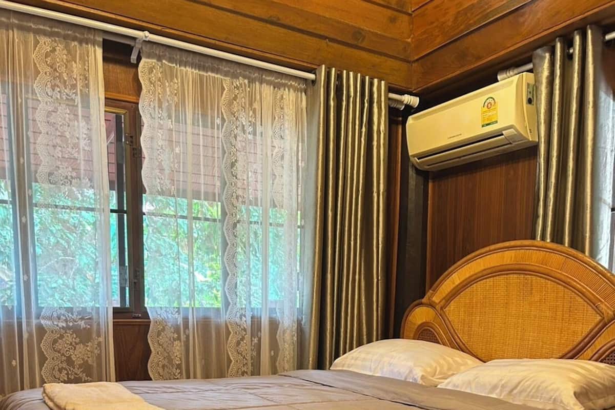 A wooden-framed room with a bed, a window with sheer curtains, and an air conditioning unit.