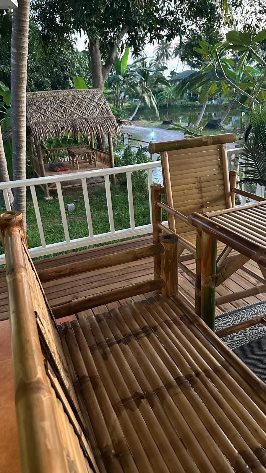 Eine Holzterrasse mit einem Bambusstuhl und einem Tisch, mit Blick auf einen Garten mit einem Gebäude mit Strohdach.