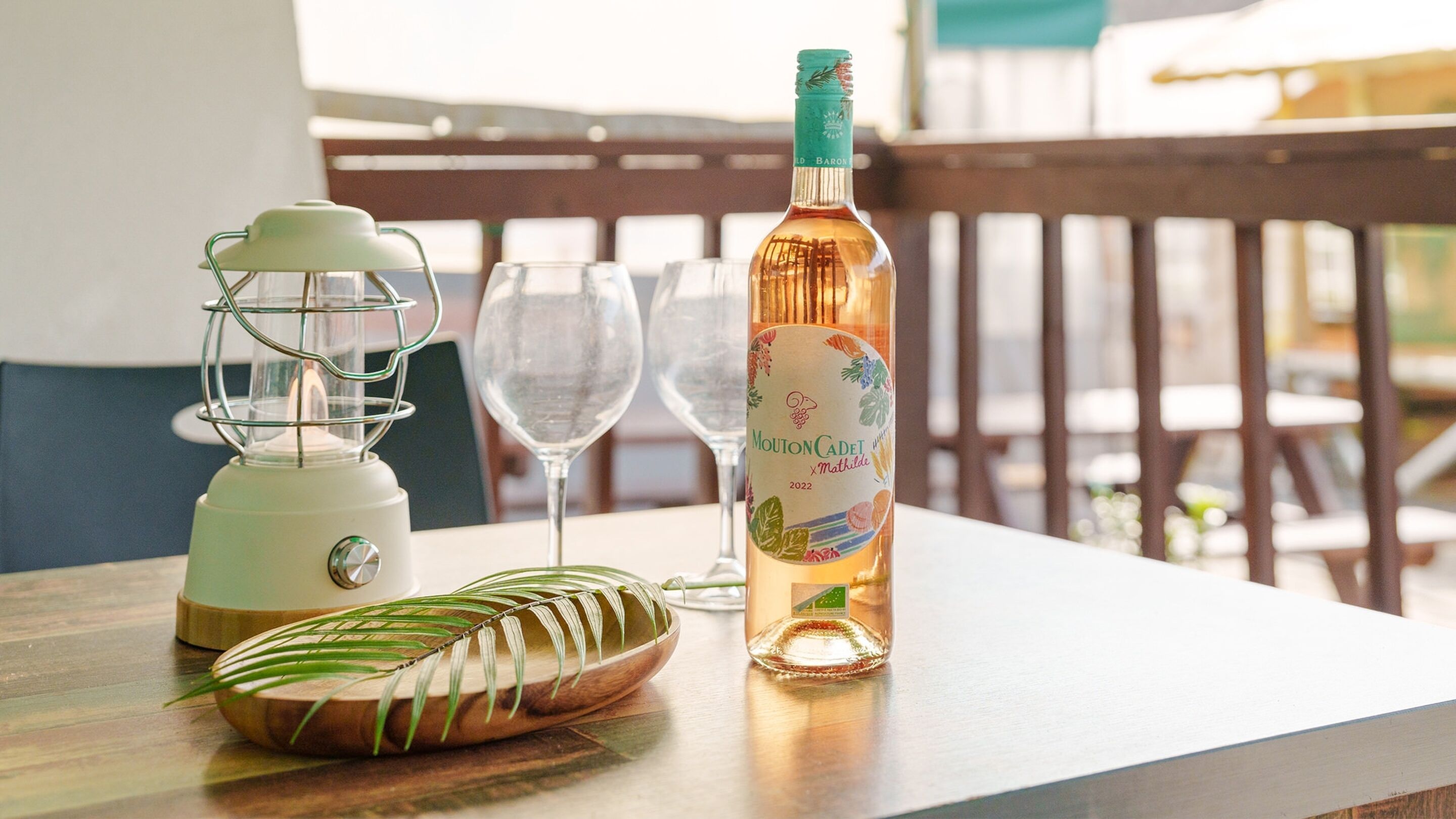 A table with a bottle of Mouton Cadet wine, a green blender, and a wooden bowl with a leaf.