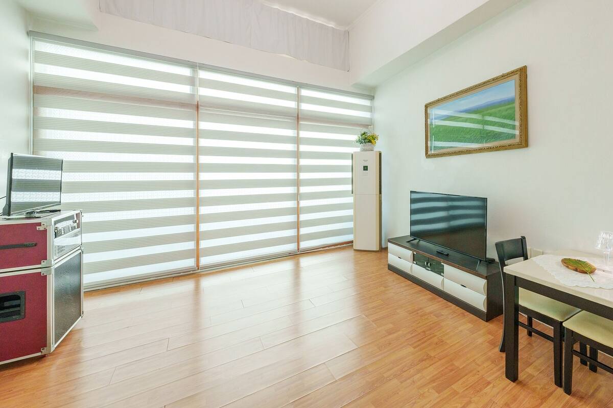 Una sala de estar moderna con una televisión de pantalla plana, piso de madera, una mesa de comedor con sillas, un refrigerador y ventanas grandes con persianas.