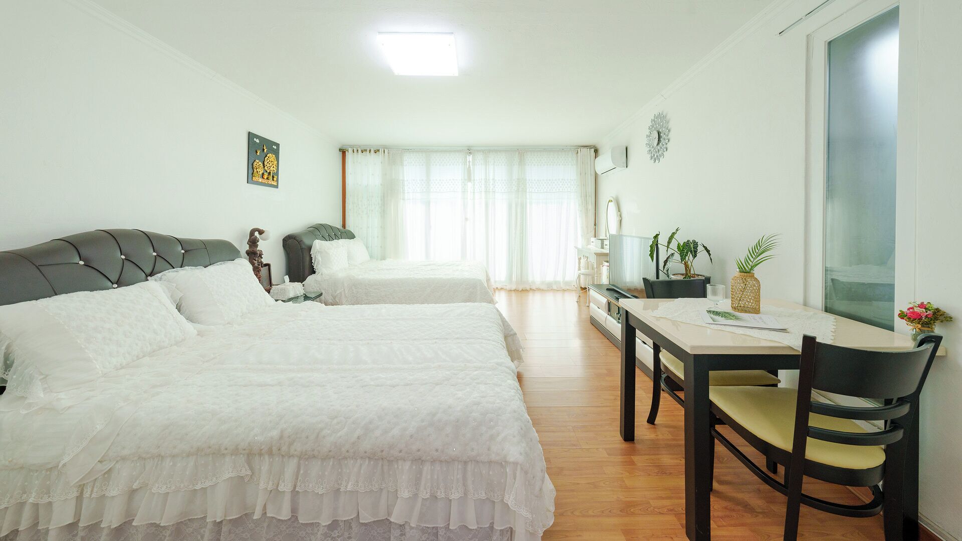 A hotel room with two beds, a dining table, chairs, a small table with a plant, and a window with curtains.