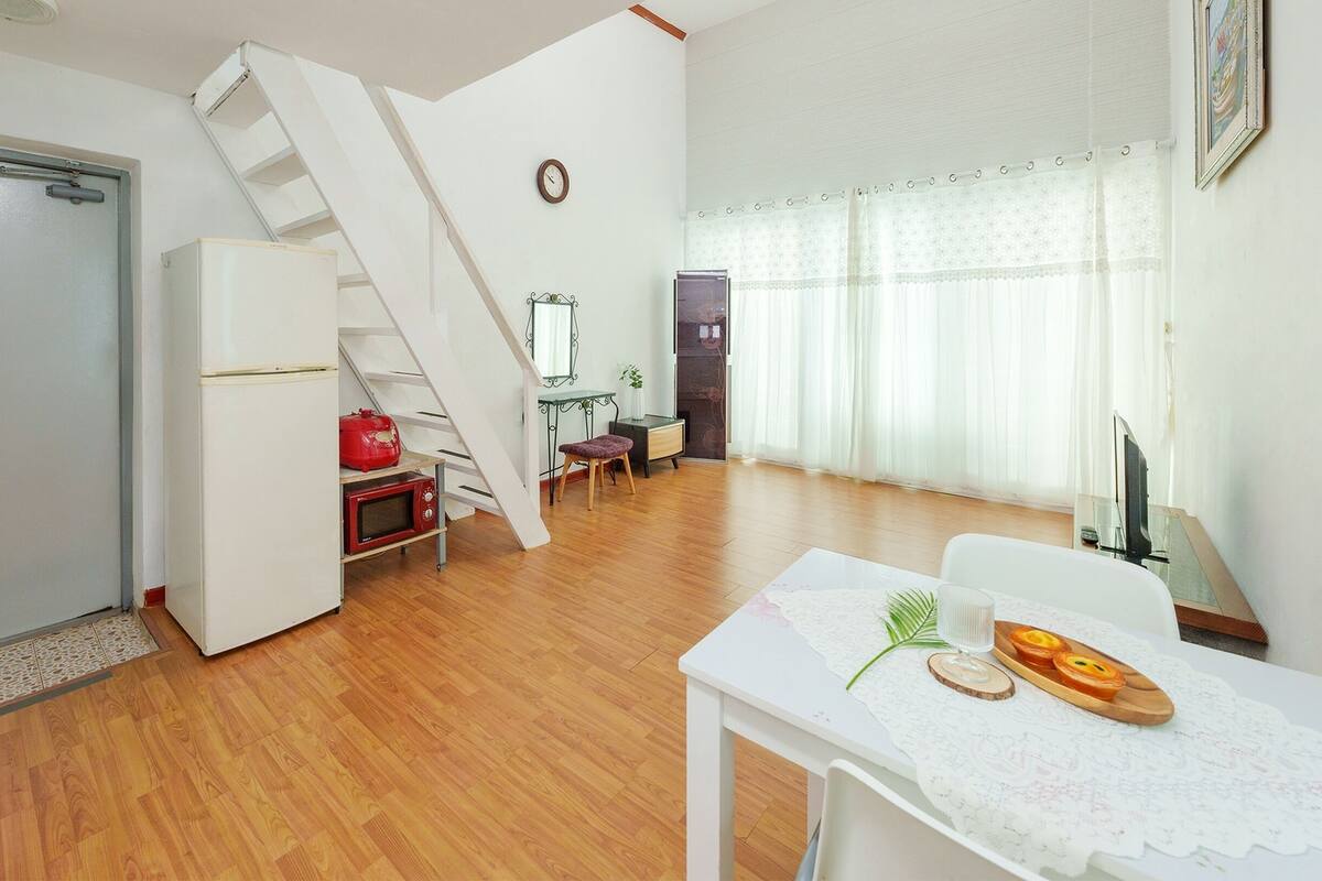 Una habitación con piso de madera, una puerta blanca, un refrigerador blanco, una mesa pequeña con un mantel individual, una tetera roja, un microondas, una silla, un taburete pequeño, un reloj y una ventana con cortinas translúcidas, además de una escalera.