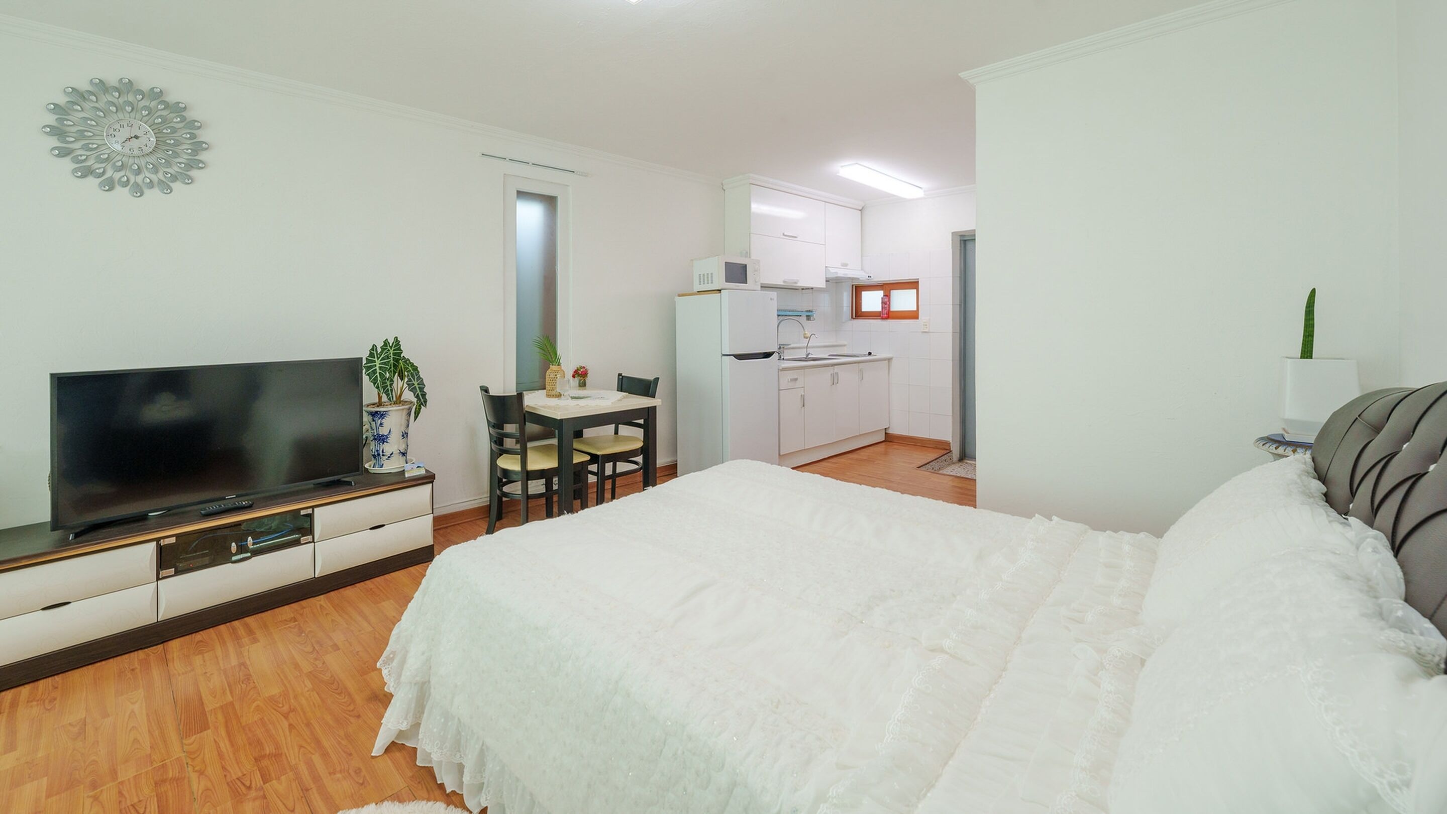A bedroom with a bed, a TV, a desk with a chair, a kitchen area, and a bathroom.