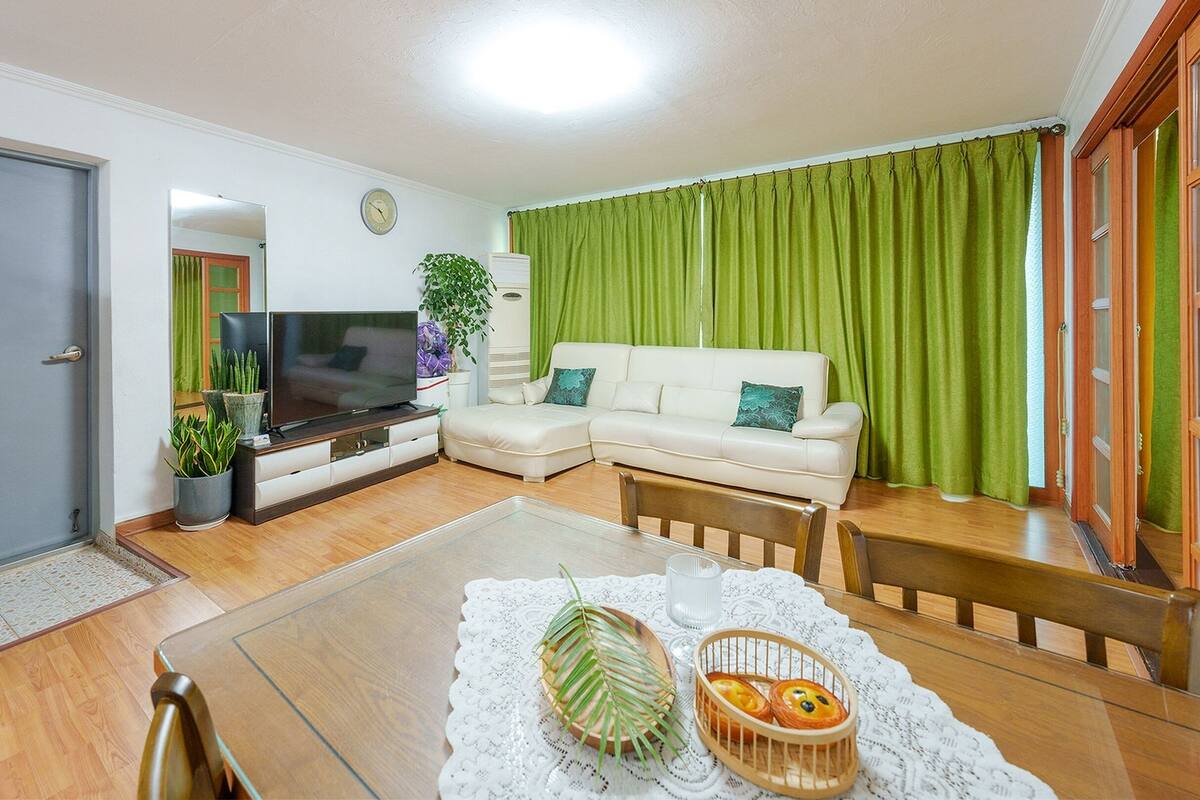 Una sala de estar con un sofá, un televisor, una mesa de comedor y un mantel individual, además de una cortina verde.