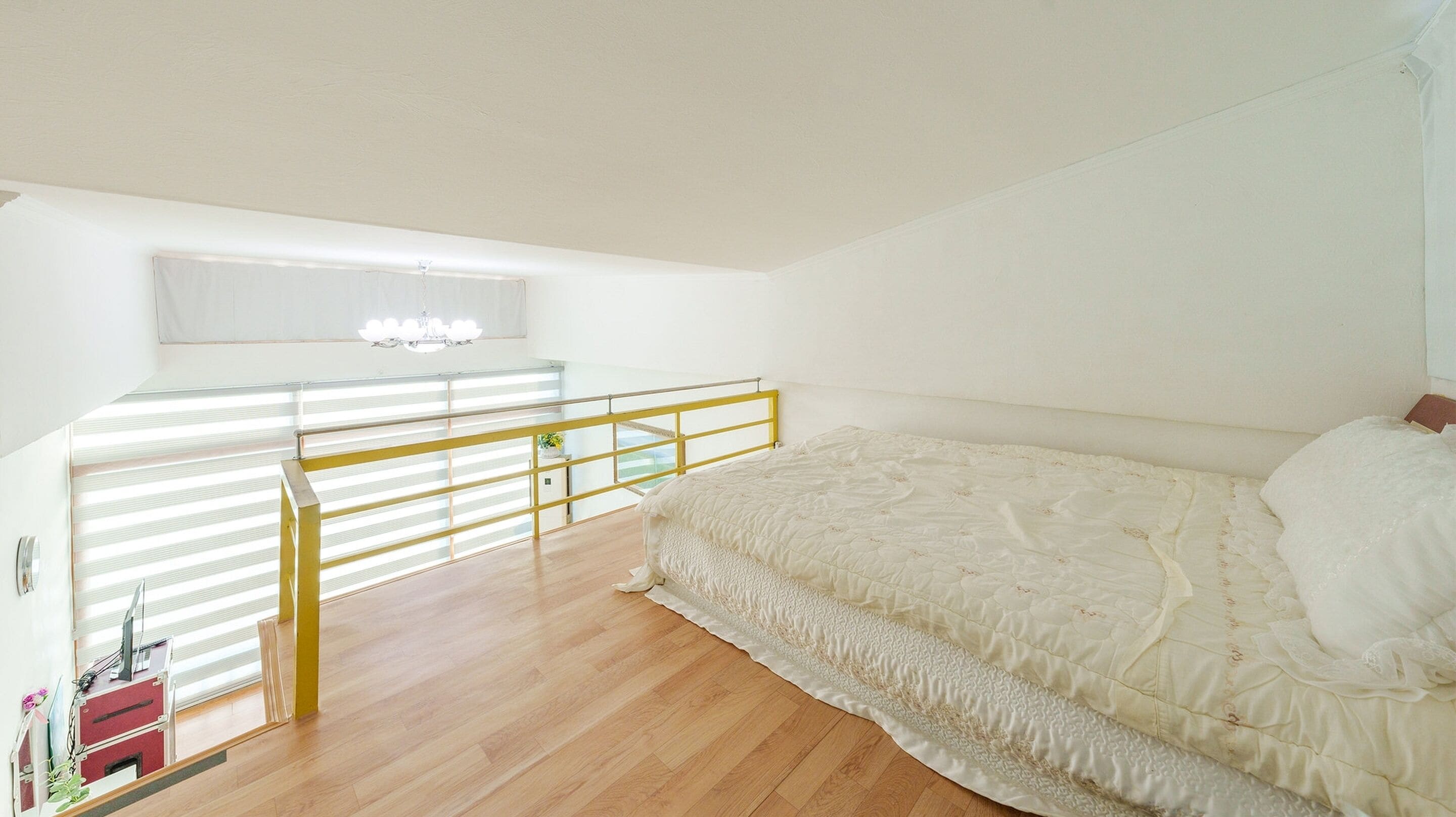 A bedroom with a bed, a window with blinds, a wooden floor, and a yellow railing.