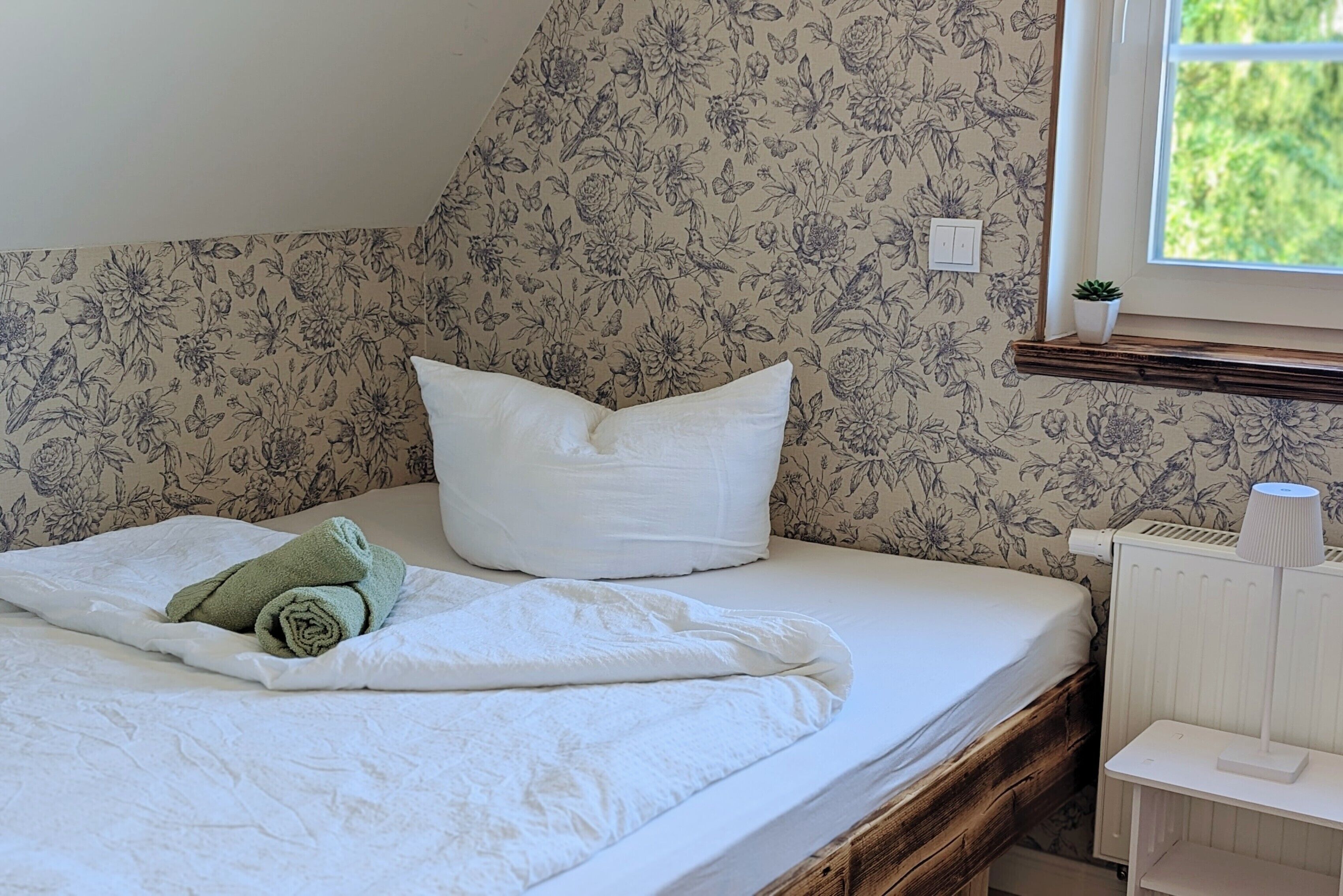 Ein Bett mit weißer Matratze, einem aufgerollten grünen Handtuch und einem weißen Kopfkissen.