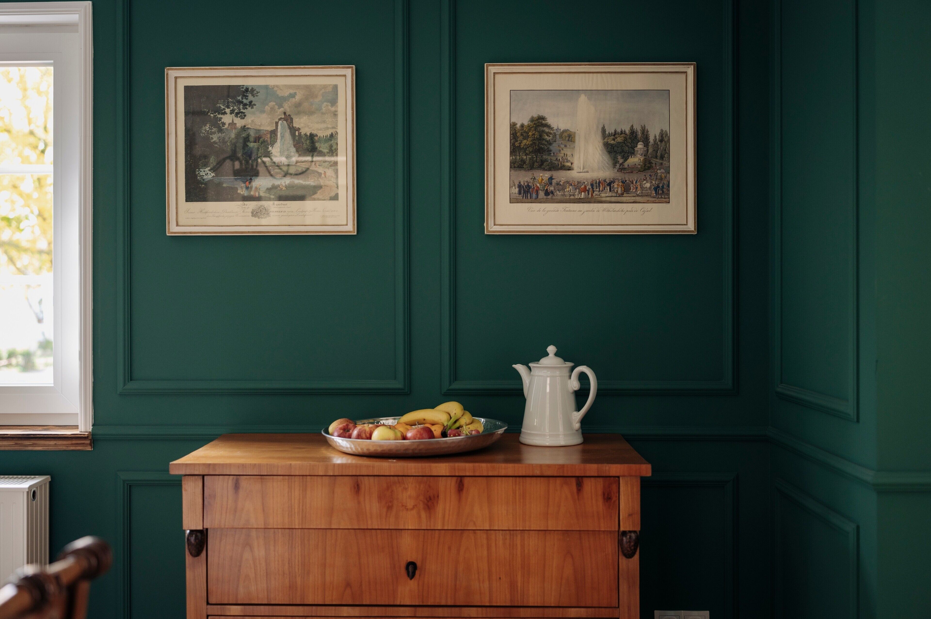 Una habitación con paredes verdes, un aparador de madera, una jarra blanca y dos cuadros enmarcados.