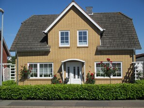 Une maison à deux étages, dotée d’un toit à pignon, d’un porche à l’avant et d’une haie bien entretenue.