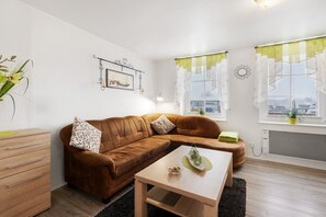 Un salon moderne avec un canapé en cuir marron, une table basse en bois et une petite table d’appoint. On y trouve une fenêtre donnant sur la ville, un radiateur et un cadre photo fixé au mur.
