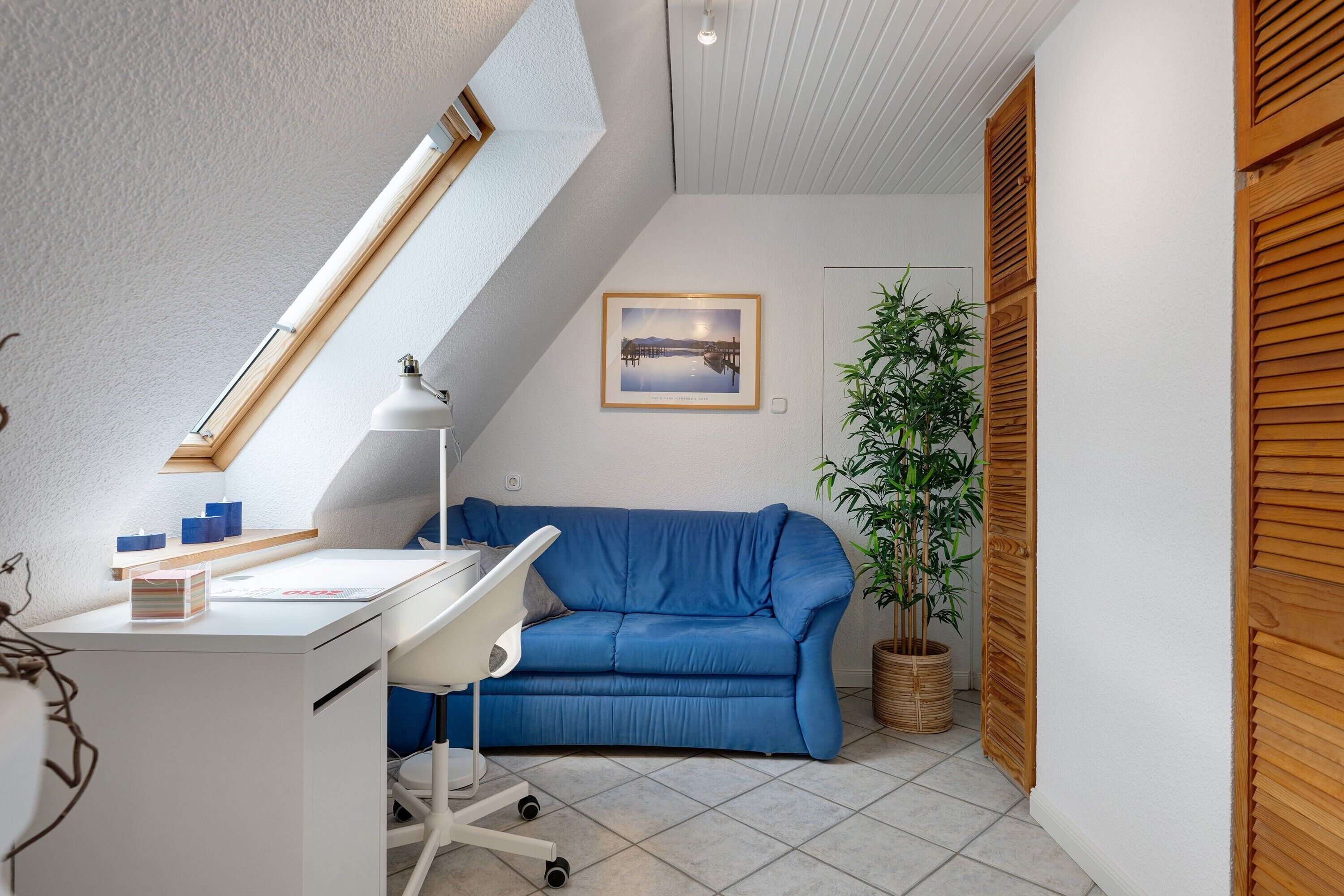 Una habitación con escritorio, sofá azul, planta en maceta, cuadro enmarco y tragaluz.