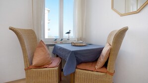 Ein Zimmer mit zwei Korbstühlen, einem Tisch mit gestreifter Tischdecke, einem Fenster mit Ausblick und einem Wandspiegel.