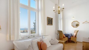Ein Zimmer mit weißem Sofa, Korbsessel, Esstisch und Meerblick durch große Fenster.