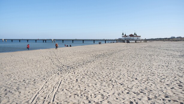 Ein weiter Sandstrand mit einem Steg, der sich ins Wasser erstreckt, ein weißes Gebäude mit rotem Dach und ein klarer, blauer Himmel.