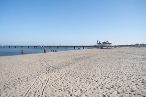 A wide sandy beach with a pier extending into the water, a white building with a red roof, and a clear blue sky.