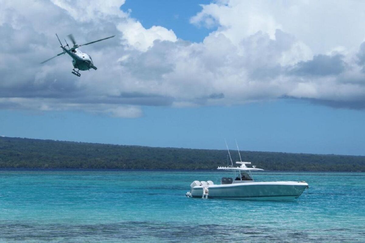 Un helicóptero vuela sobre una masa de agua, con un bote y una zona boscosa al fondo.