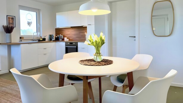 A modern kitchen with white cabinets, a dining table with white chairs, and a vase with flowers on the table. - Apartment 'Boddenbude Lubmin' with Wi-Fi (Lubmin)