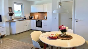 A modern kitchen with white cabinets, a wooden countertop, a built-in oven, and a dining area with a round table and chairs. - Apartment 'Boddenbude Lubmin' with Wi-Fi (Lubmin)