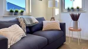 A modern living room with a grey sofa, decorative pillows, a floor lamp, and potted plants on the windowsill. - Apartment 'Boddenbude Lubmin' with Wi-Fi (Lubmin)