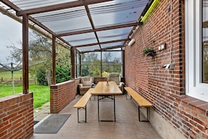 A covered patio with a brick wall, a wooden table, benches, and a chair. - Holiday Home 'Hamdorf' with Private Terrace, Shared Garden and Wi-Fi (Hamdorf)