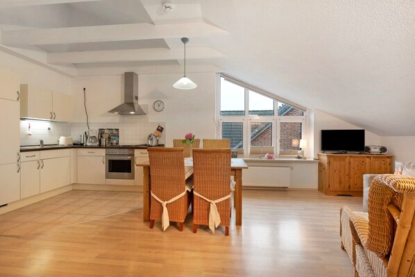 A modern kitchen with white cabinets, a dining area with wicker chairs, a wooden dining table, and a TV in the living room.