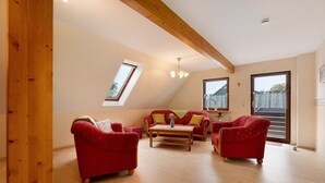 A room with a sloped ceiling, wooden beams, a red sofa set, a wooden coffee table, and a large window. - Holiday Apartment Norderwurth (Büsumer Deichhausen)