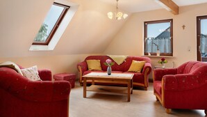A loft-style room with a sloped ceiling, a wooden ceiling beam, a red sofa, a wooden coffee table, and a window with a view of a house. - Holiday Apartment Norderwurth (Büsumer Deichhausen)
