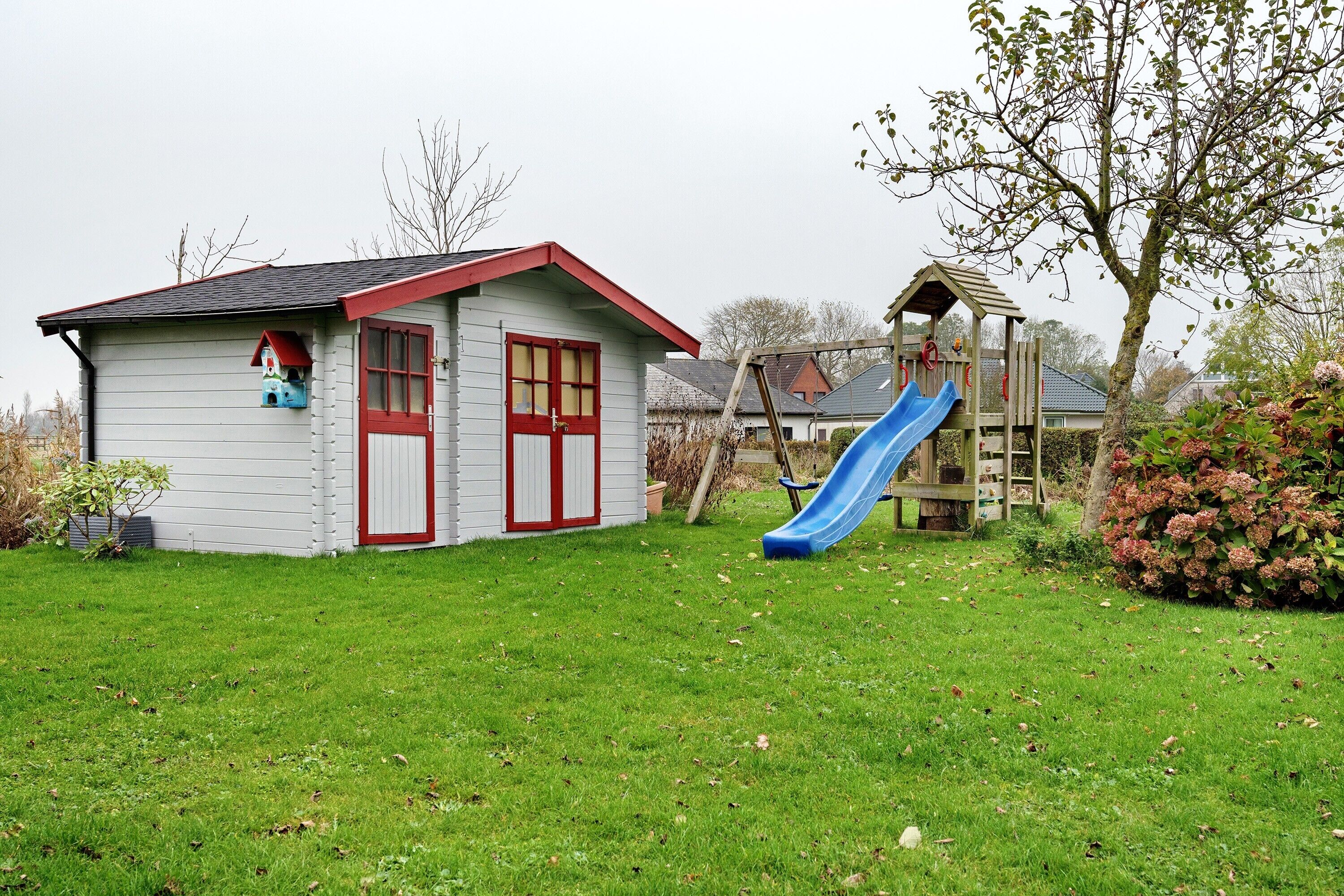Ein kleines Haus mit roter Tür und Fenstern, einer blauen Rutsche und einem hölzernen Spielhäuschen.