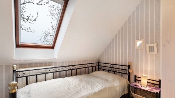 Une chambre avec un plafond en pente, un lit avec une tête de lit, une table de chevet avec une lampe et une fenêtre donnant sur des branches d’arbres nues.