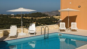 Un espace piscine avec des chaises longues blanches et des parasols, un mur en pierre, et une vue sur la mer et les collines en arrière-plan.